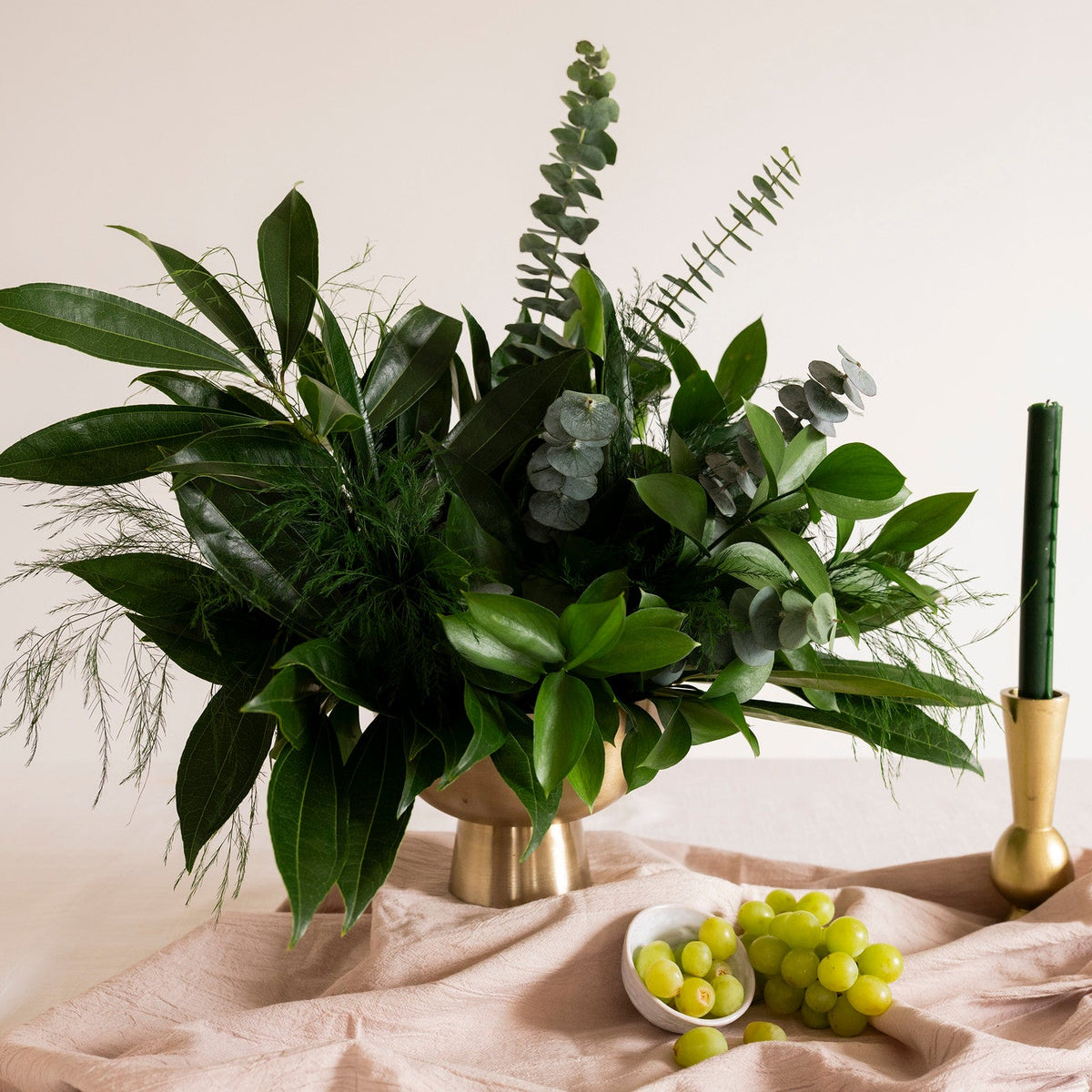 A green floral arrangement in a metallic gold vase, styled on a draped blush pink fabric surface with a small bowl of green grapes and a gold candlestick holder against a warm cream background.