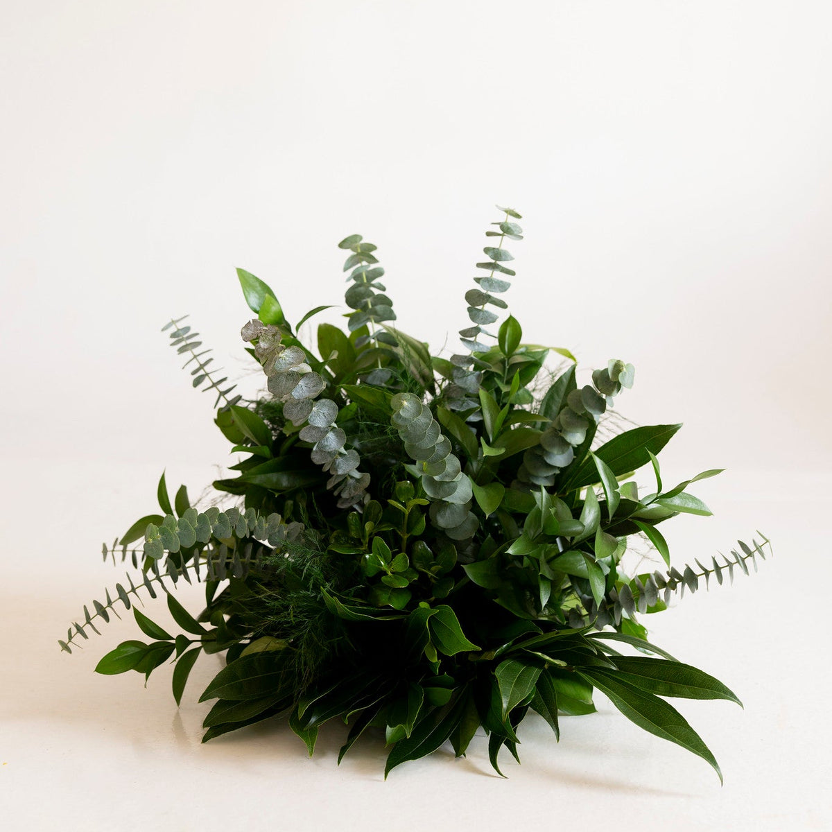 A lush green artificial floral arrangement featuring eucalyptus, ferns, and other foliage, displayed on a clean, solid off-white background.