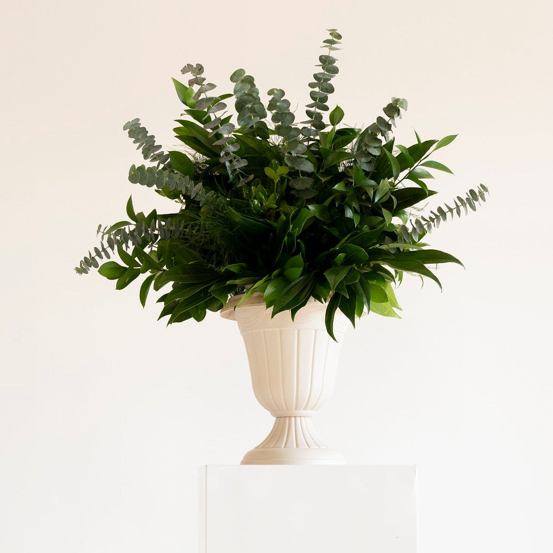 A green leafy floral arrangement in a white urn-shaped vase, displayed on a white pedestal against a plain white background.