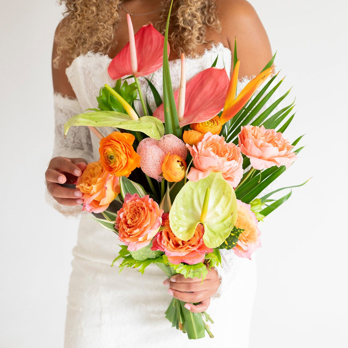 A woman in a white strapless wedding dress holds a vibrant bridal bouquet featuring coral roses, orange ranunculus, pink anthuriums, and green foliage against a plain white background.
