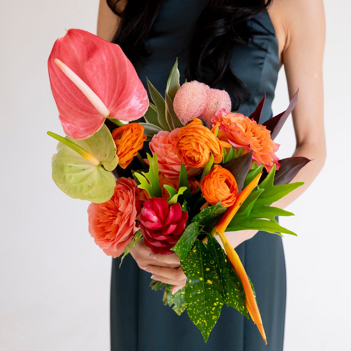 A woman in a dark teal sleeveless dress holds a vibrant bouquet of tropical flowers, including pink and orange anthuriums, roses, and green leaves, against a plain white background.