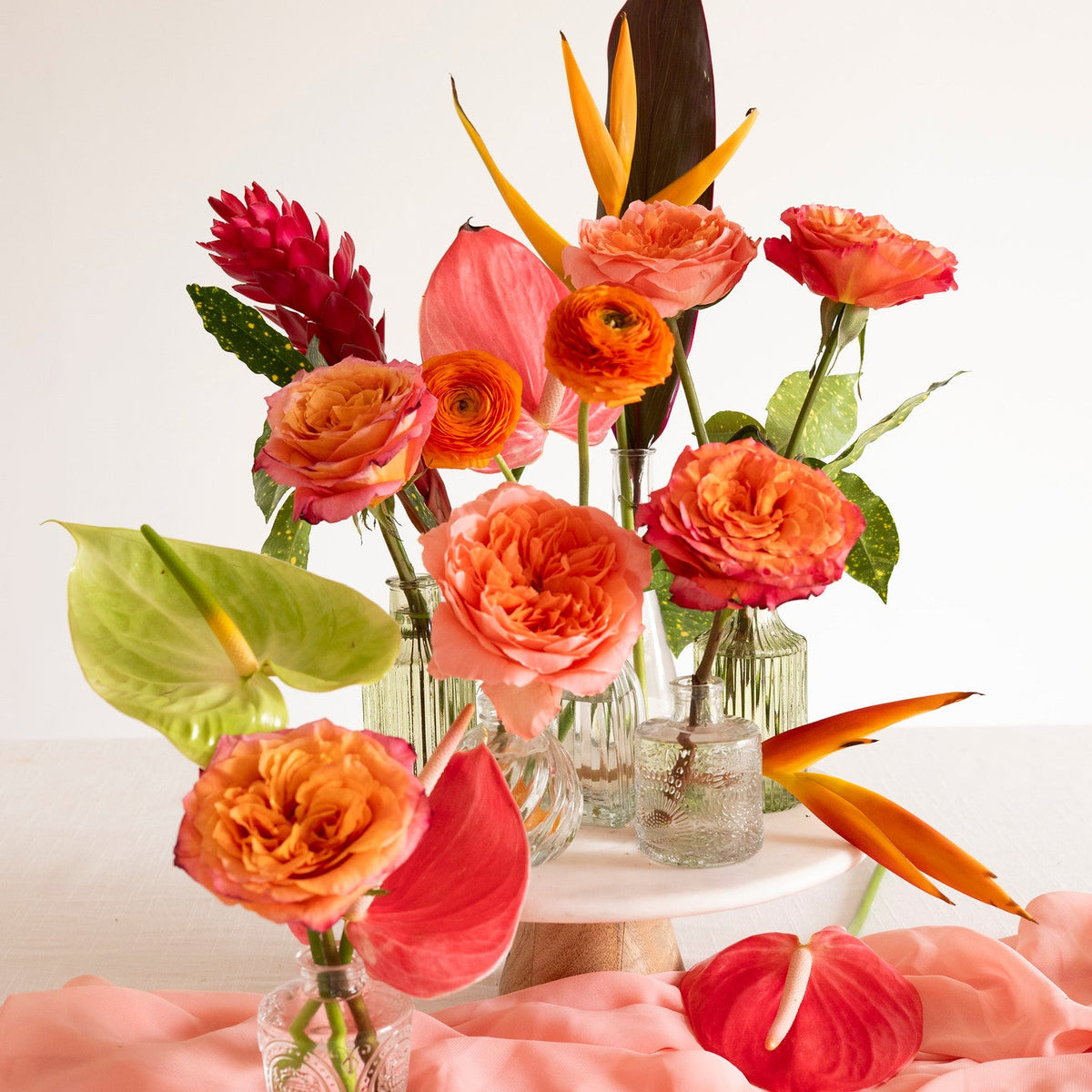 A vibrant tropical floral arrangement featuring orange roses, pink and red anthuriums, green leaves, and tropical bird of paradise flowers in clear glass vases, styled on a draped pink fabric surface against a clean white background.