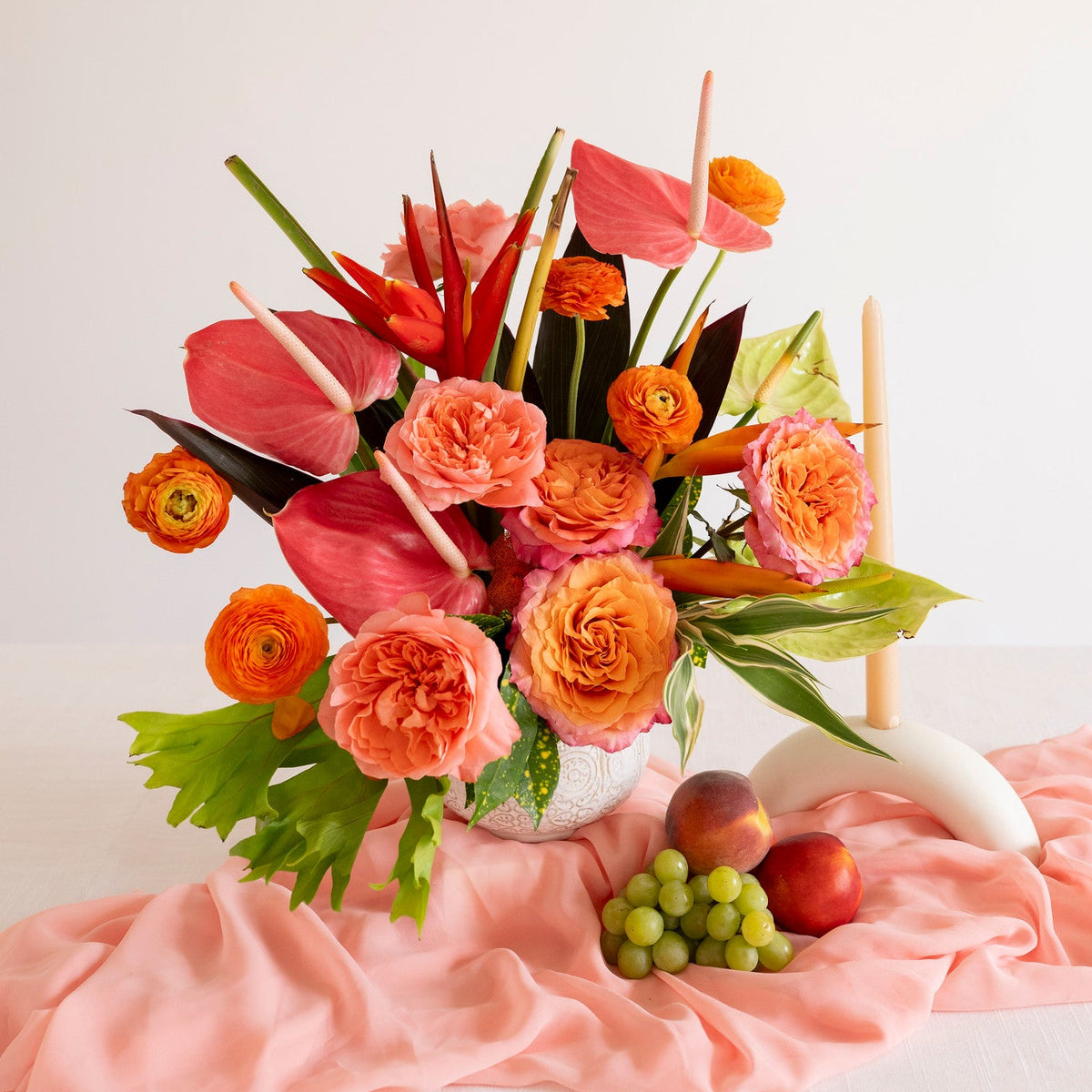 A vibrant tropical floral arrangement featuring pink and orange flowers with green leaves, styled in a white vase on a draped pink fabric surface with small fruits and a white candle holder.