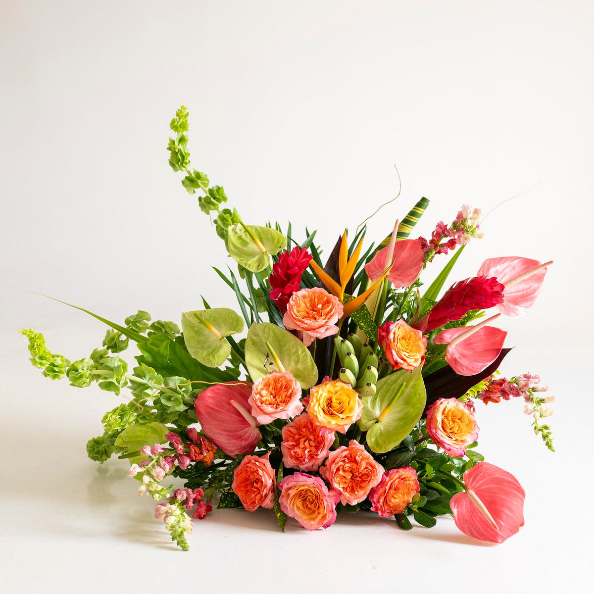 A vibrant floral arrangement featuring pink and orange roses, red anthuriums, green foliage, and tropical bird of paradise flowers, photographed on a clean white studio background.