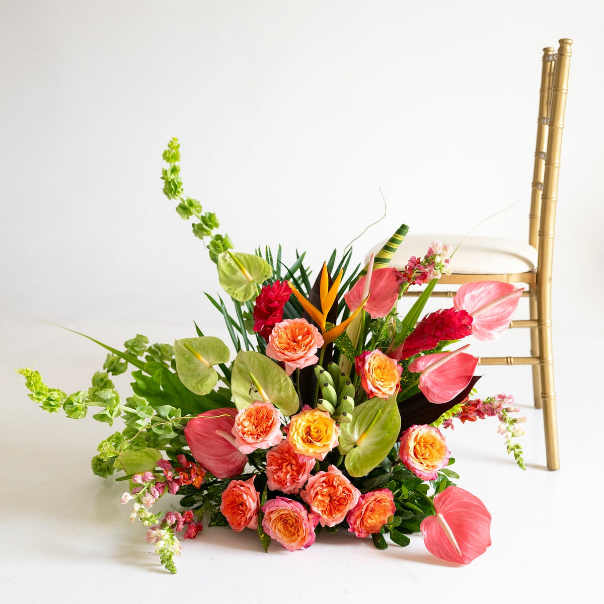 A vibrant tropical floral arrangement featuring pink and orange roses, green foliage, and tropical flowers, styled next to a gold Chiavari chair on a clean white background.