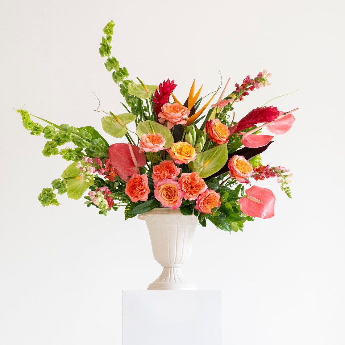 A vibrant floral arrangement featuring pink, red, and orange flowers with green foliage, presented in a white classical-style urn on a white pedestal against a clean white background.