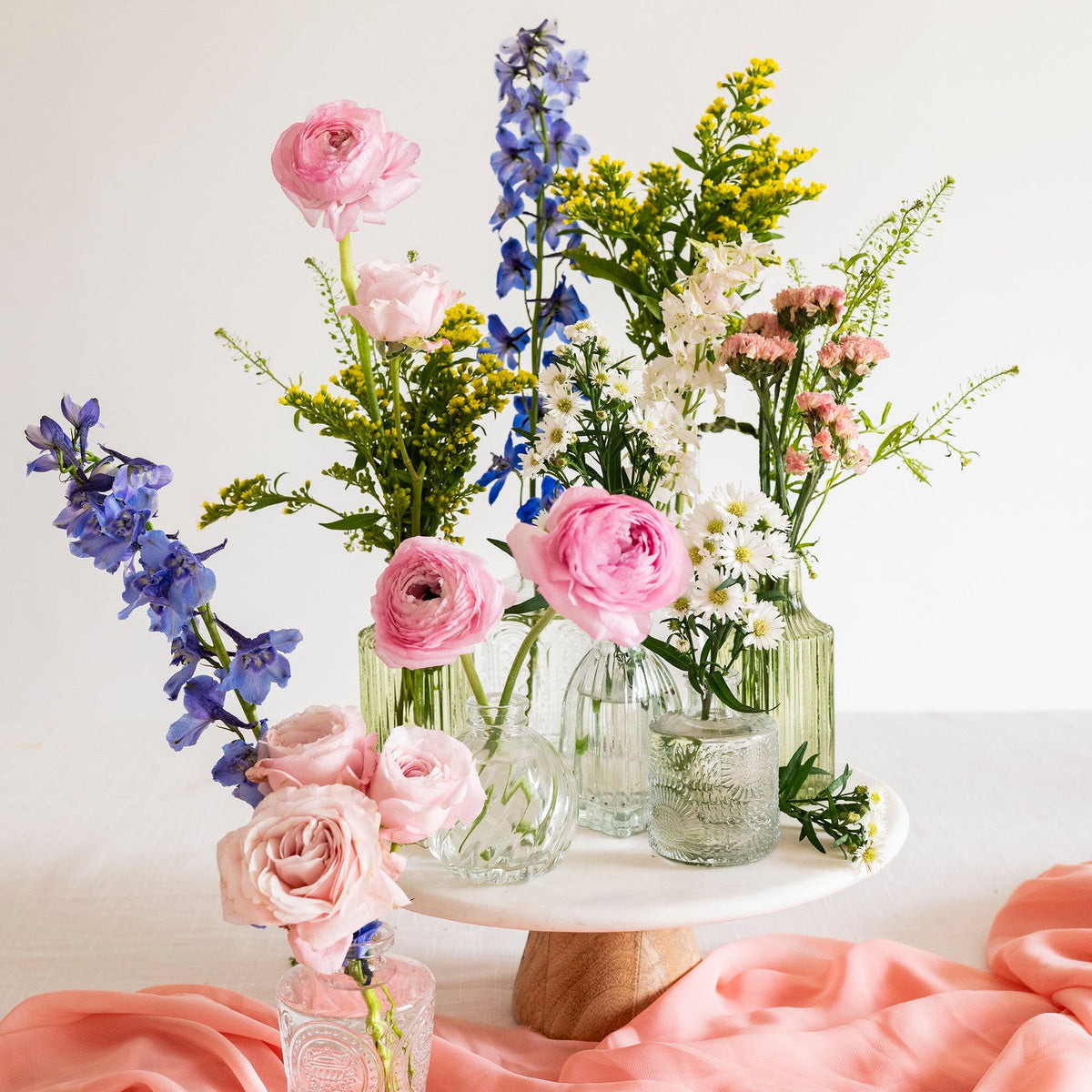 A styled floral arrangement in clear glass vases on a white pedestal stand, with pink roses, blue delphiniums, white daisies, and green foliage, set on a white surface with draped peach fabric in the foreground.
