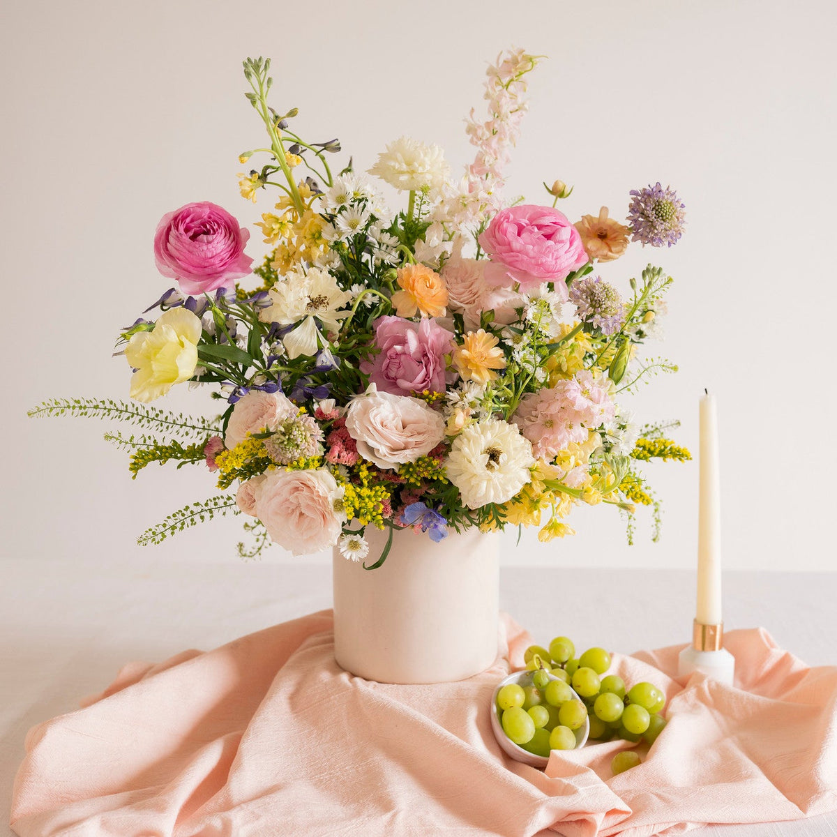 A pastel floral arrangement in a white vase on a draped peach fabric, with a small bowl of green grapes and a lit taper candle beside it against a soft off-white background.