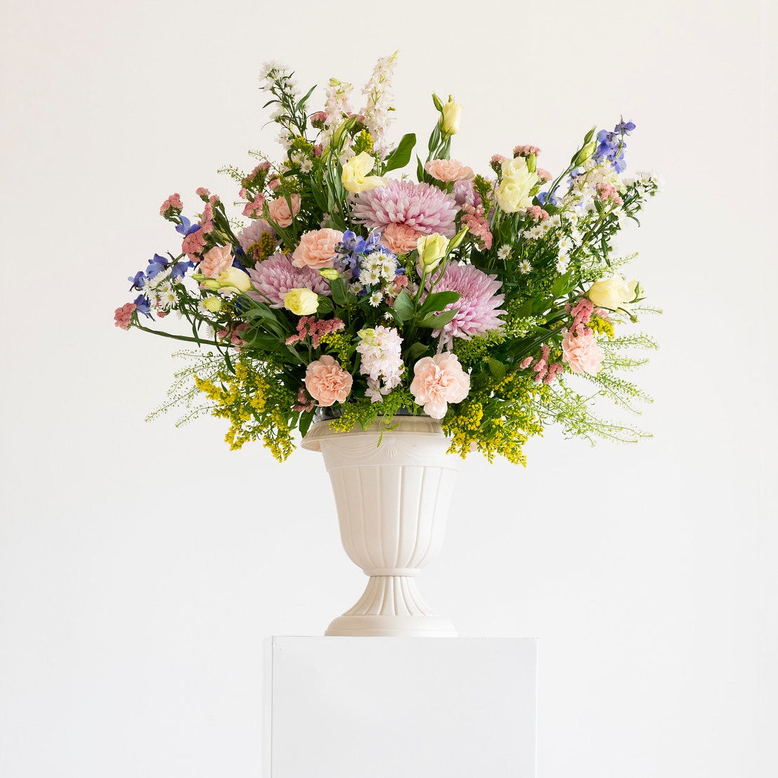 A wildflower flower bouquet in a white urn-shaped vase, displayed on a white pedestal against a clean white background.